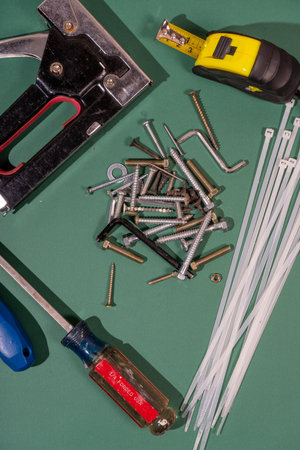 Diy tools including cable ties, screws, screwdriver, tape measure and staple gun lying on green background, ready to be used for home maintenanceの写真素材