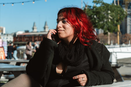 Stylish woman with bright red hair is sitting at an outdoor table, making a phone call on a sunny dayの写真素材