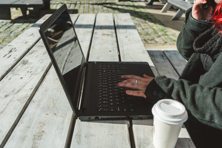 Freelancer woman typing on laptop keyboard and talking on phone while working remotely outdoorsの写真素材