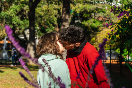 Romantic couple sharing a tender kiss in a vibrant park setting, surrounded by colorful flowers and treesの写真素材