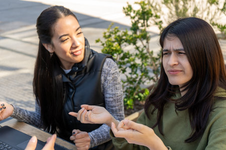 Two young women are discussing a project on a laptop at an outdoor table, one gesturing and looking...