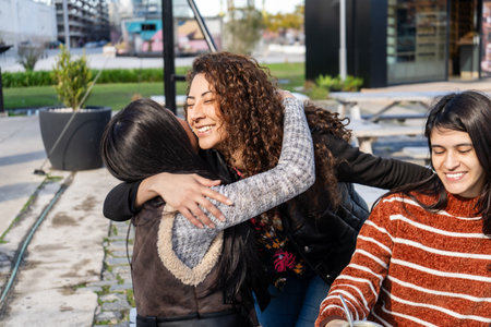 Three cheerful young women embrace at a table outside a cafe, enjoying friendship and connectionの写真素材