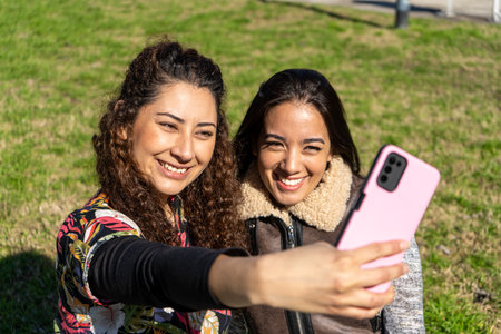 Two happy female friends taking a selfie with a smartphone in a park, enjoying a sunny day outdoorsの写真素材