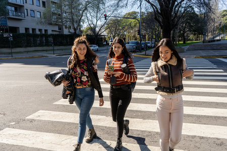 Three female students are walking and chatting on a pedestrian crossing in the city during a sunny dayの写真素材