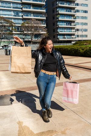 Excited woman walking and waving while holding shopping bags in a cityの写真素材