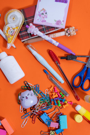 Flat lay of stationery on an orange surface, featuring pens, scissors, glue, paper clips, and thumbtacks, perfect for educational projectsの写真素材