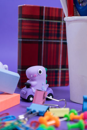 Playful miniature dinosaur driving purple toy car surrounded by colorful stationery, erasers, paper clips, and push pins on a vibrant purple backdropの写真素材