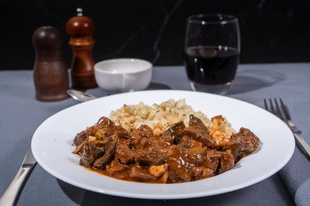 Hungarian goulash with spaetzle and a glass of red wine served on a white plate, a hearty and traditional mealの写真素材