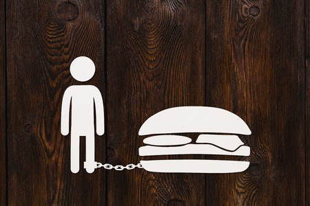 Paper man chained to burger, fastfood or overeating concept, dark wooden background, abstract conceptual imageの写真素材