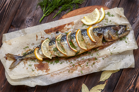 Whole baked mackerel or scomber fish with lemon and greens on paper, dark wooden background, top viewの写真素材