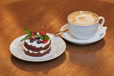 Round cake with cream, berries and cup of latte coffee on white plateの写真素材