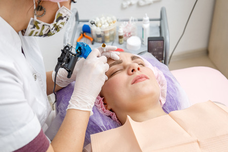 Permanent makeup, tattooing of eyebrows. Cosmetologist in white gloves applying make up with machine for woman in beauty salonの写真素材