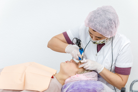 Permanent makeup, tattooing of eyebrows. Cosmetologist in white gloves applying make up with machine for woman in beauty salonの写真素材