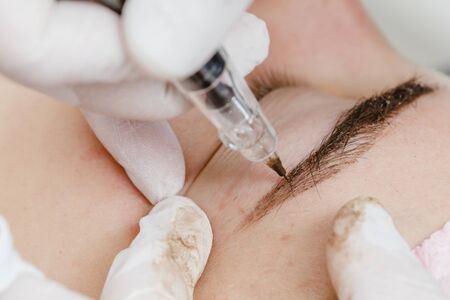 Permanent makeup, tattooing of eyebrows. Cosmetologist in white gloves applying make up with machine...