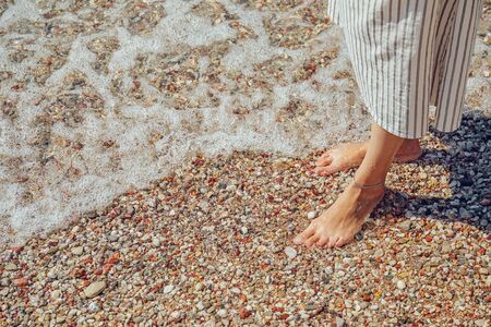 Beautiful woman legs on pebble beach with foamy transparent sea waterの写真素材