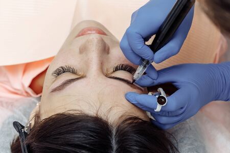 Permanent makeup, tattooing of eyebrows. Cosmetologist in white gloves applying make up with machine for woman in beauty salonの写真素材