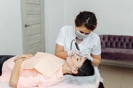 Permanent makeup, tattooing of eyebrows. Cosmetologist in white gloves applying make up with machine for woman in beauty salonの写真素材