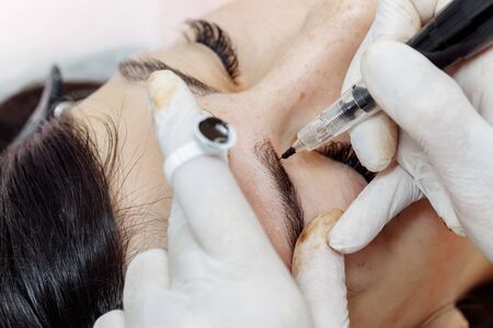 Permanent makeup, tattooing of eyebrows. Cosmetologist in white gloves applying make up with machine for woman in beauty salonの写真素材