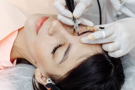 Permanent makeup, tattooing of eyebrows. Cosmetologist in white gloves applying make up with machine for woman in beauty salonの写真素材