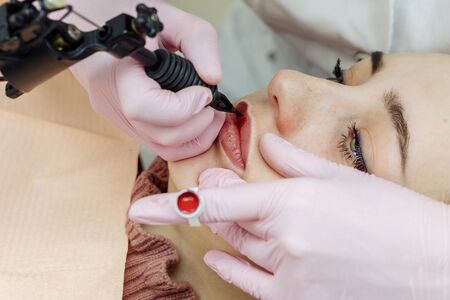Woman having permanent makeup on lips in beauty salon. Cosmetologist in gloves applying tattoo with machine, macro viewの写真素材