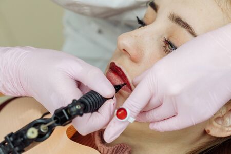 Woman having permanent makeup on lips in beauty salon. Cosmetologist in gloves applying tattoo with machine, macro viewの写真素材