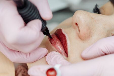 Woman having permanent makeup on lips in beauty salon. Cosmetologist in gloves applying tattoo with machine, macro viewの写真素材