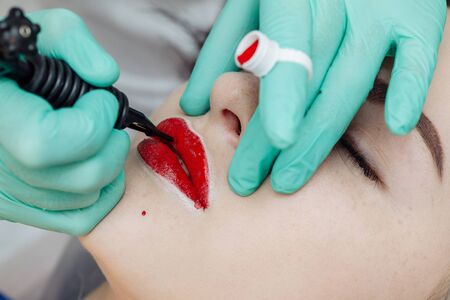 Woman having permanent makeup on lips in beauty salon. Cosmetologist in gloves applying tattoo with machine, macro viewの写真素材