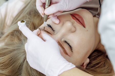 Permanent makeup, tattooing of eyebrows. Cosmetologist in white gloves applying make up with machine for woman in beauty salonの写真素材