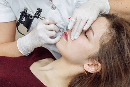 Woman having permanent makeup on lips in beauty salon. Cosmetologist in gloves applying tattoo with machine, macro viewの写真素材