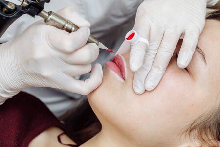 Woman having permanent makeup on lips in beauty salon. Cosmetologist in gloves applying tattoo with machine, macro viewの写真素材