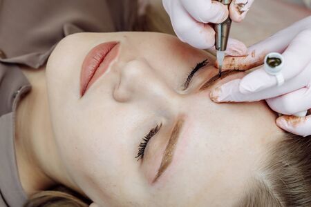Permanent makeup, tattooing of eyebrows. Cosmetologist in white gloves applying make up with machine for woman in beauty salonの写真素材