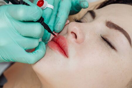 Woman having permanent makeup on lips in beauty salon. Cosmetologist in gloves applying tattoo with machine, macro viewの写真素材