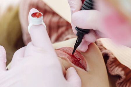 Woman having permanent makeup on lips in beauty salon. Cosmetologist in gloves applying tattoo with machine, macro viewの写真素材