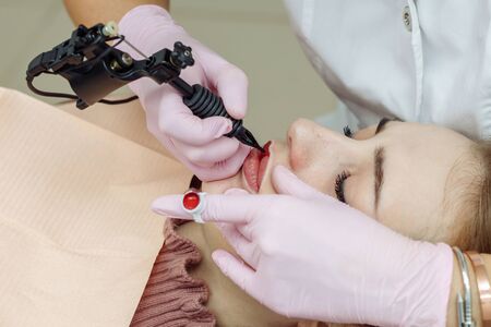 Woman having permanent makeup on lips in beauty salon. Cosmetologist in gloves applying tattoo with machine, macro viewの写真素材