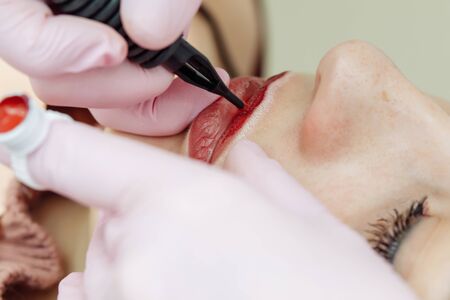 Woman having permanent makeup on lips in beauty salon. Cosmetologist in gloves applying tattoo with machine, macro viewの写真素材