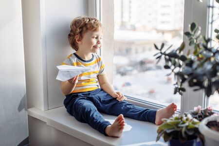 Child boy sitting near window with paper plane and looking. Stay home concept, Coronavirus Covid-19 quarantineの写真素材