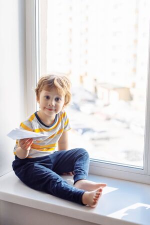 Child boy sitting near window with paper plane and looking. Stay home concept, Coronavirus Covid-19 quarantineの写真素材