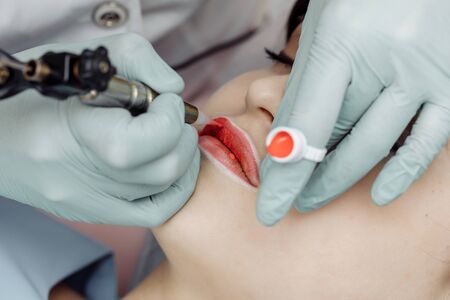 Woman having permanent makeup on lips in beauty salon. Cosmetologist in gloves applying tattoo with machine, macro viewの写真素材
