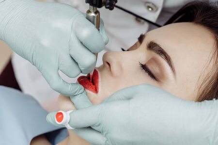 Woman having permanent makeup on lips in beauty salon. Cosmetologist in gloves applying tattoo with machine, macro viewの写真素材