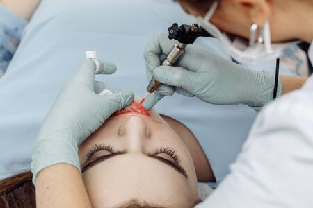 Woman having permanent makeup on lips in beauty salon. Cosmetologist in gloves applying tattoo with machine, macro viewの写真素材