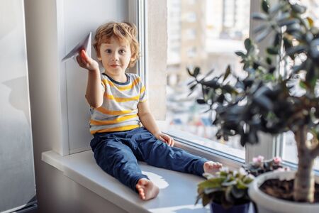 Child boy sitting near window with paper plane and looking. Stay home concept, Coronavirus Covid-19 quarantineの写真素材