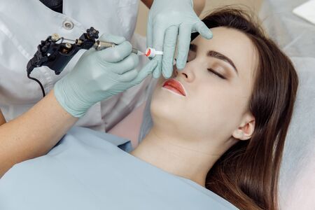 Woman having permanent makeup on lips in beauty salon. Cosmetologist in gloves applying tattoo with machine, macro viewの写真素材