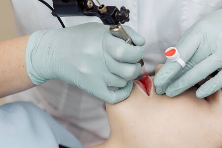 Woman having permanent makeup on lips in beauty salon. Cosmetologist in gloves applying tattoo with machine, macro viewの写真素材