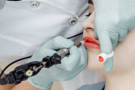 Woman having permanent makeup on lips in beauty salon. Cosmetologist in gloves applying tattoo with machine, macro viewの写真素材