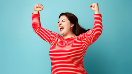 Portrait of a happy young woman celebrating success with arms raised on blue background. Enthusiastic overweight Caucasian woman emotionally raises her hands. Happy young woman in red striped shirt celebrating success.の素材