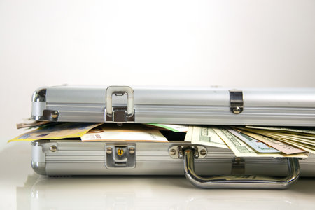 A close-up of an open silver aluminum case with cash spilling out from inside. The case is on a white surface with the cash slightly overlapping the edge.の写真素材