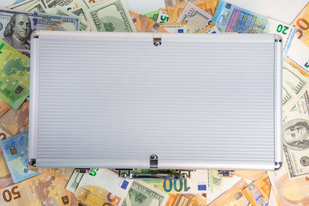 A silver briefcase is closed and lying on a white surface surrounded by US dollar bills and Euro...