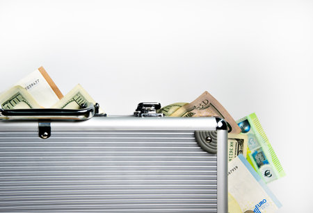 A silver suitcase with a slightly open lid, revealing several dollar bills and one euro bill peeking out from the top.の写真素材