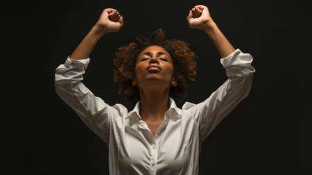 A woman expresses joy with her arms raised high, her curly hair catching the light. She is wearing a white shirt and is surrounded by a dark background, symbolizing freedom.の素材