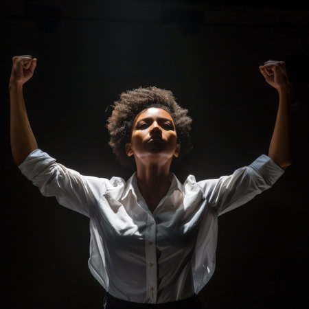A confident woman stands tall with her fists raised in triumph, illuminated by strong overhead lighting, conveying a powerful message of empowerment and strength.の素材